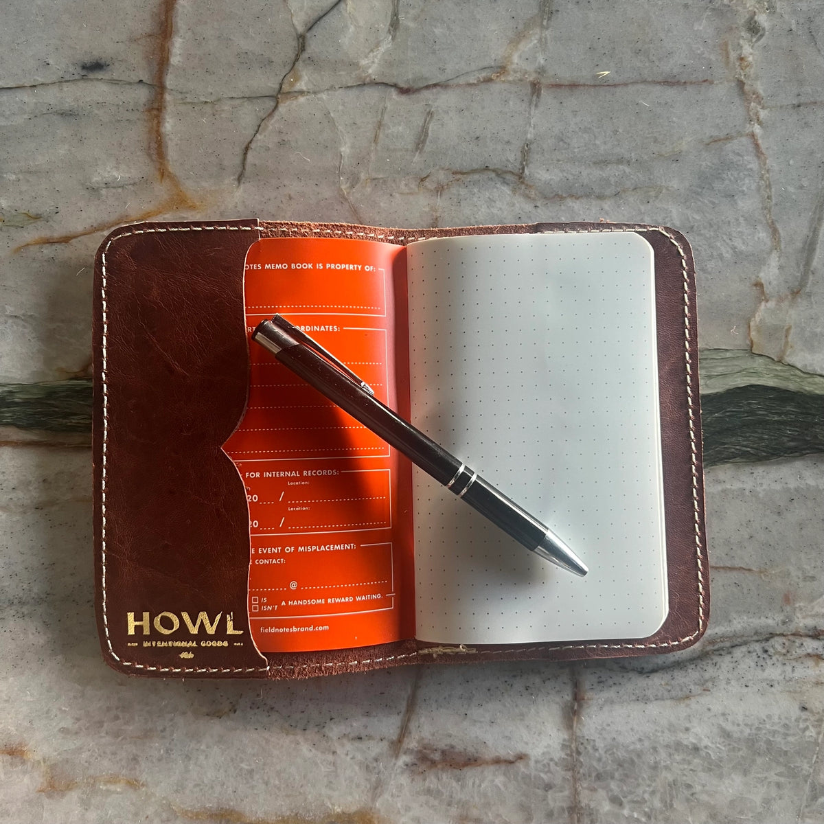 Brown leather notebook with white pages and a pen on a stone surface, featuring 'HOWL' branding.