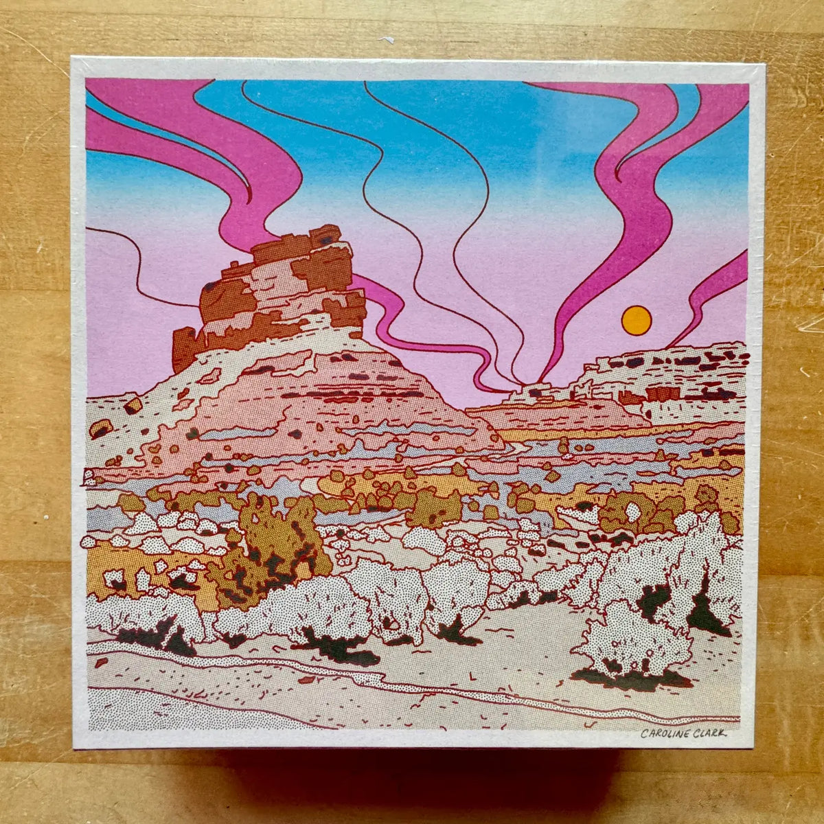 Colorful desert landscape illustration with pink and blue smoke-like patterns on a wooden surface.
