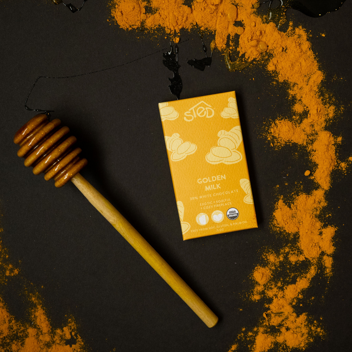 A bar of white chocolate with turmeric powder, labeled 'Golden Milk Bar', presented next to a honey dipper, all placed on a surface with a golden spice sprinkled on it.