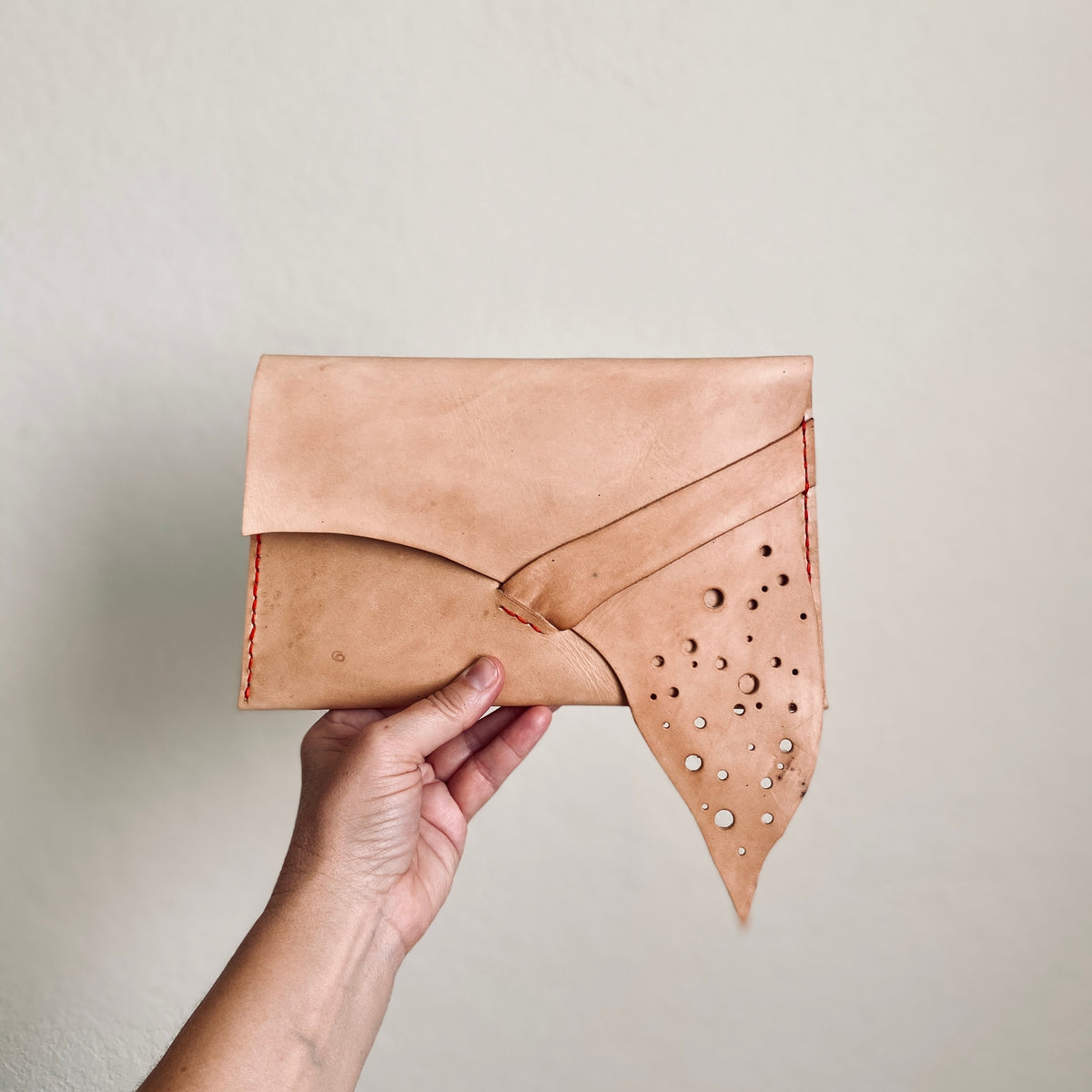 Handsewn Leather clutch with asymetrical flap that tucks under a leather strap to close and is stitched in contrasting color held by a white hand model