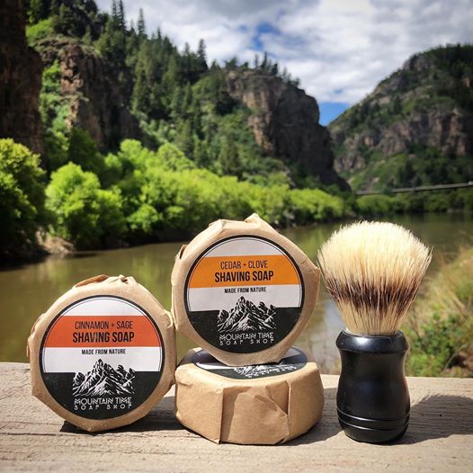 A shaving soap bar with a label reading 'CEDAR + SAGE SHAVING SOAP', a synthetic shaving brush, and a black mug, all placed on a wooden surface with a scenic mountainous background.