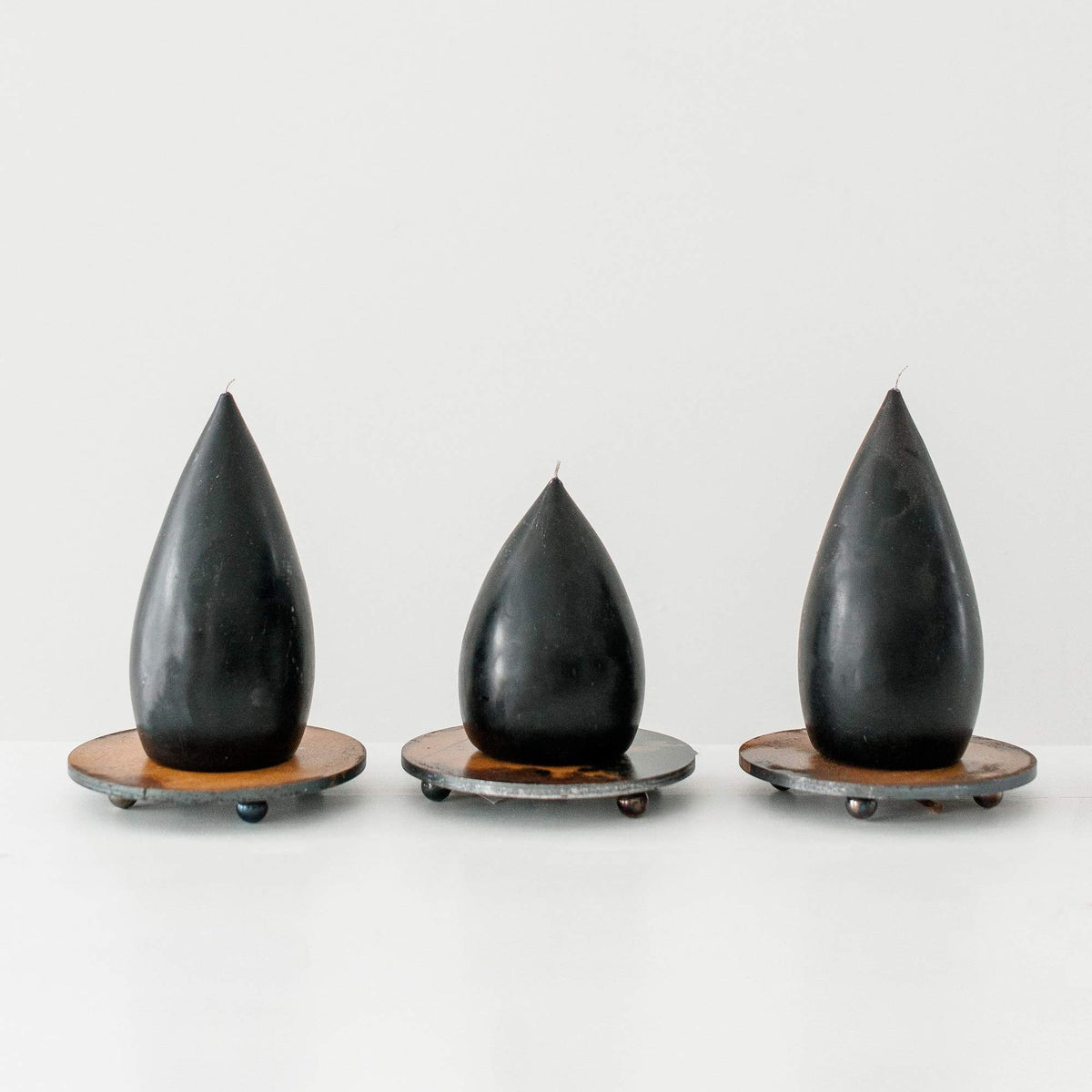 Three teardrop shaped black candles on patina steel plate candle holders on a white surface and background