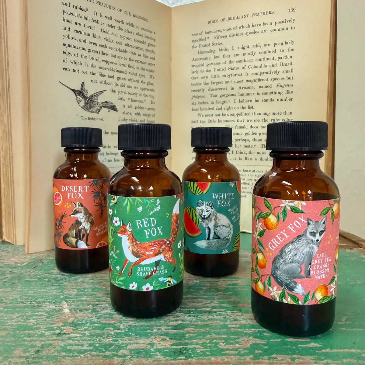 Four bottles with animal-themed labels on a wooden surface with an open book in the background.