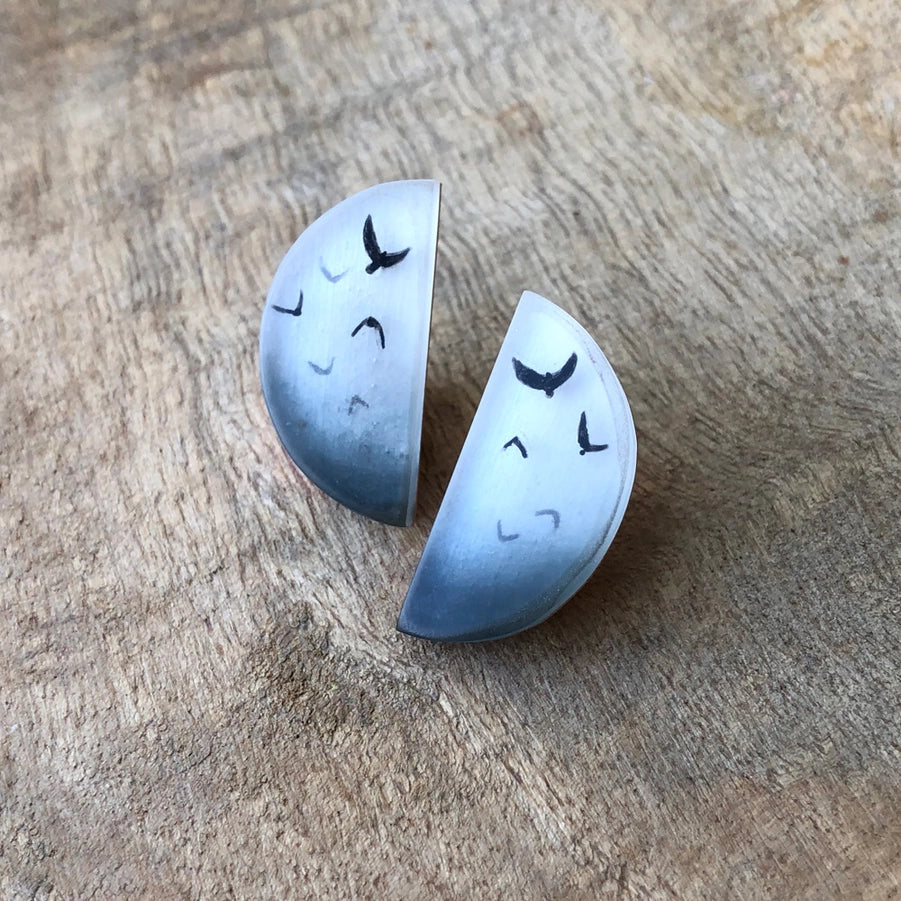 Two ceramic earrings shaped like half moons with bird illustrations on a wooden surface.