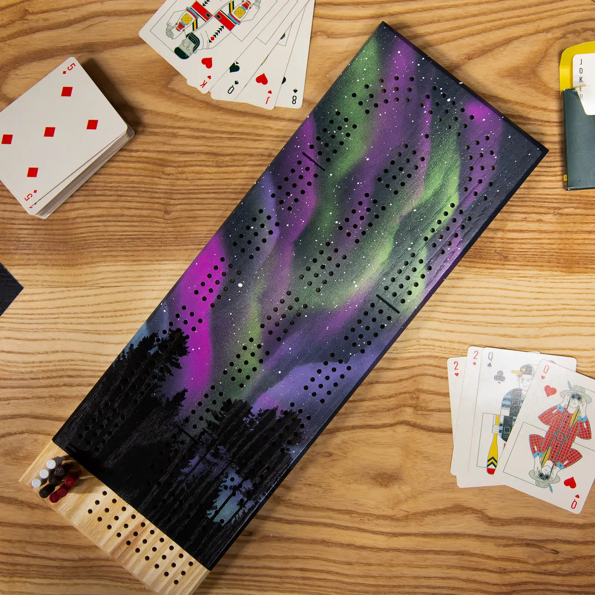 Cribbage board with Northern Lights design on a wooden table with playing cards.