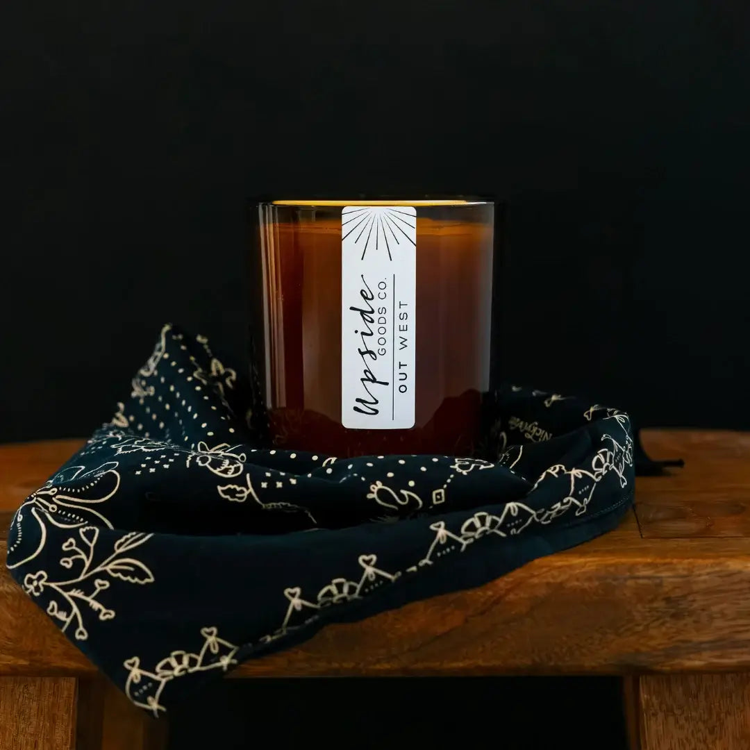 An amber glass jar candle with a white vertical label reading 'Upfide Goods Co. Out West' sits on a dark navy bandana with cream embroidery, placed on a wooden surface against a black background.