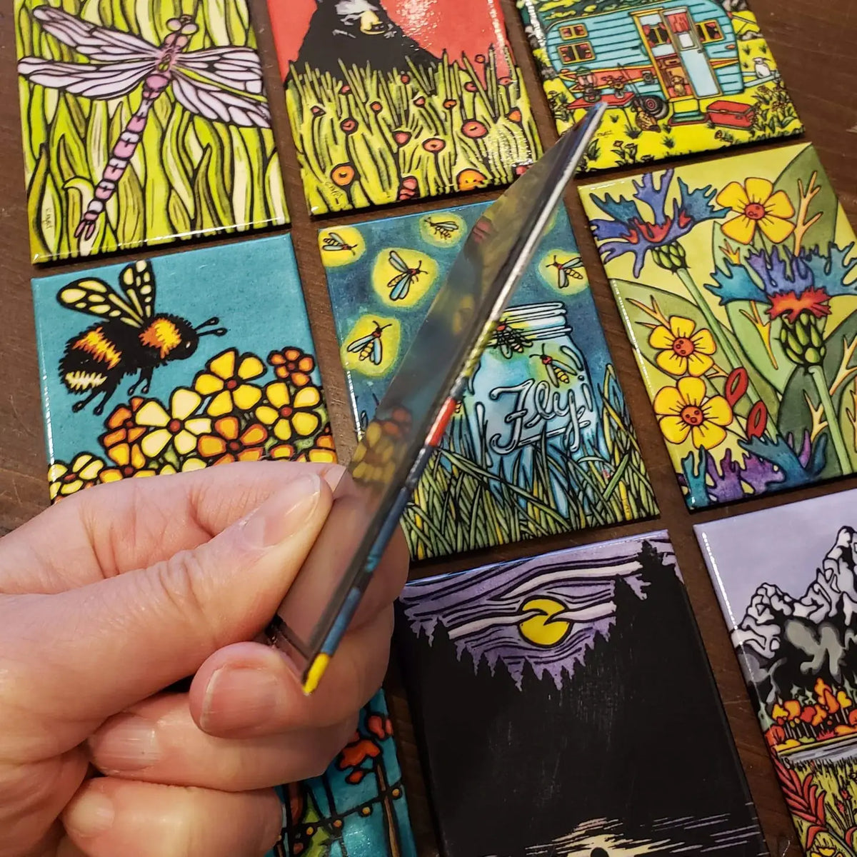 Colorful mosaic of individual magnets with various designs, including a bee and flowers. One magnet being flipped over to show side and back. 