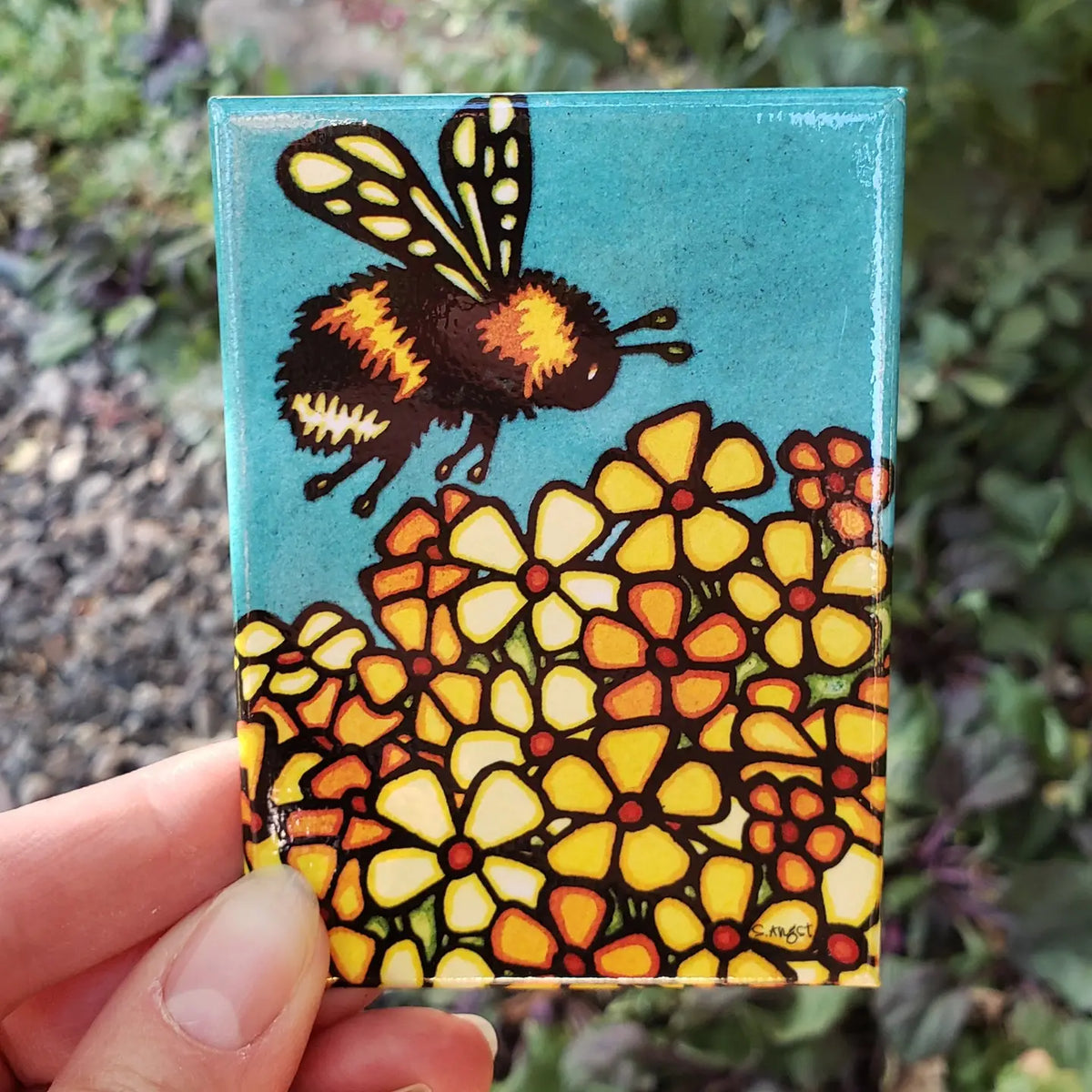 Small rectangular magnet with artwork of a butterfly on flowers held by a hand with a natural background.