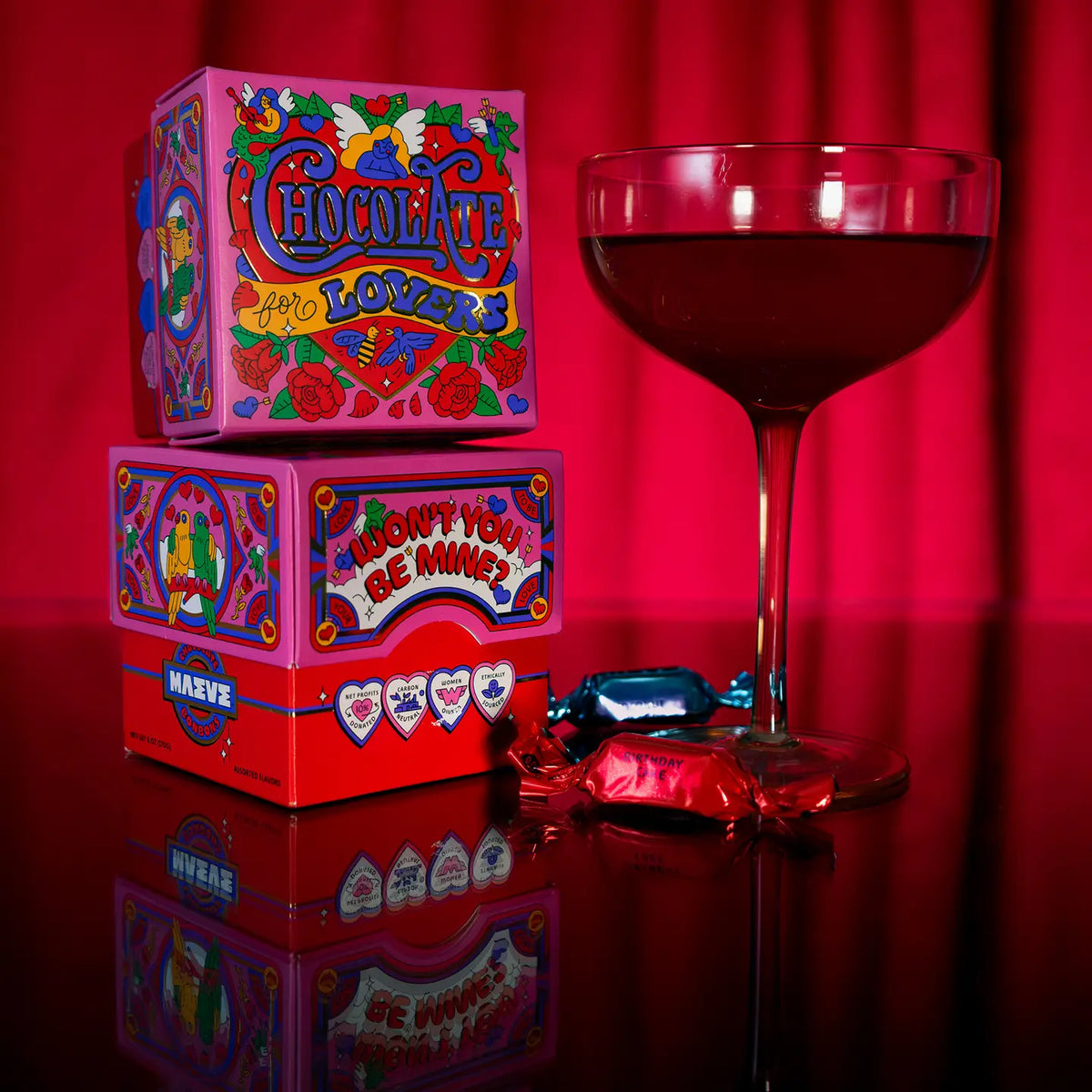 Two colorful boxes labeled 'Chocolate for Lovers' and 'Won't You Be Mine?' next to a glass of red wine on a red background.