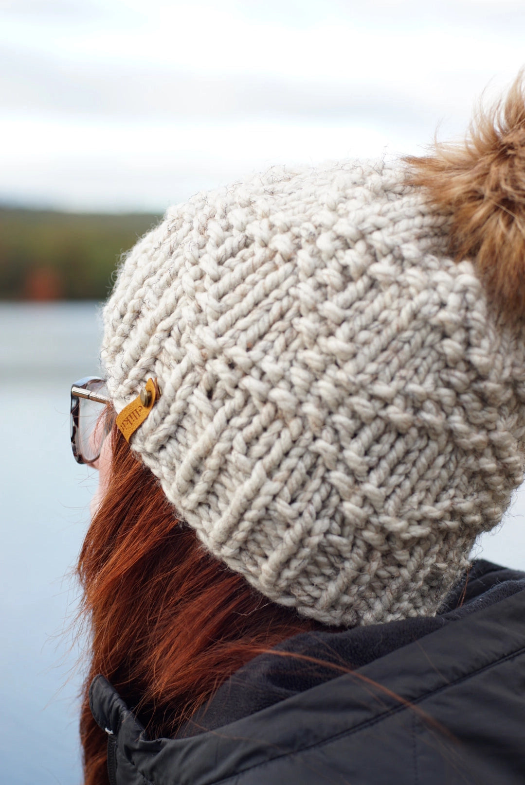 Person wearing a knitted beige beanie with a pom-pom, standing by a body of water.
