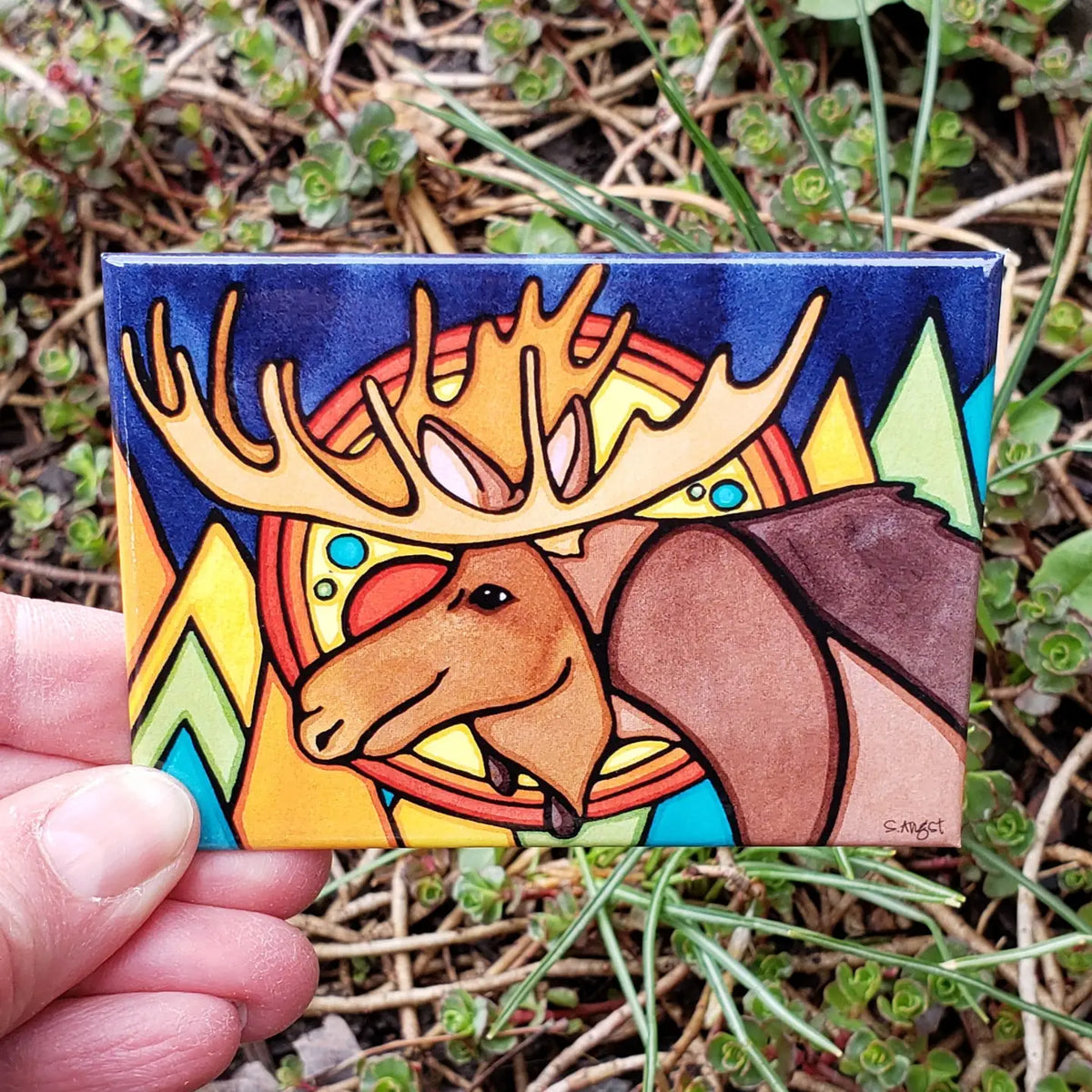 Colorful artwork magnet of a moose with a rainbow and mountain design held in front of greenery.