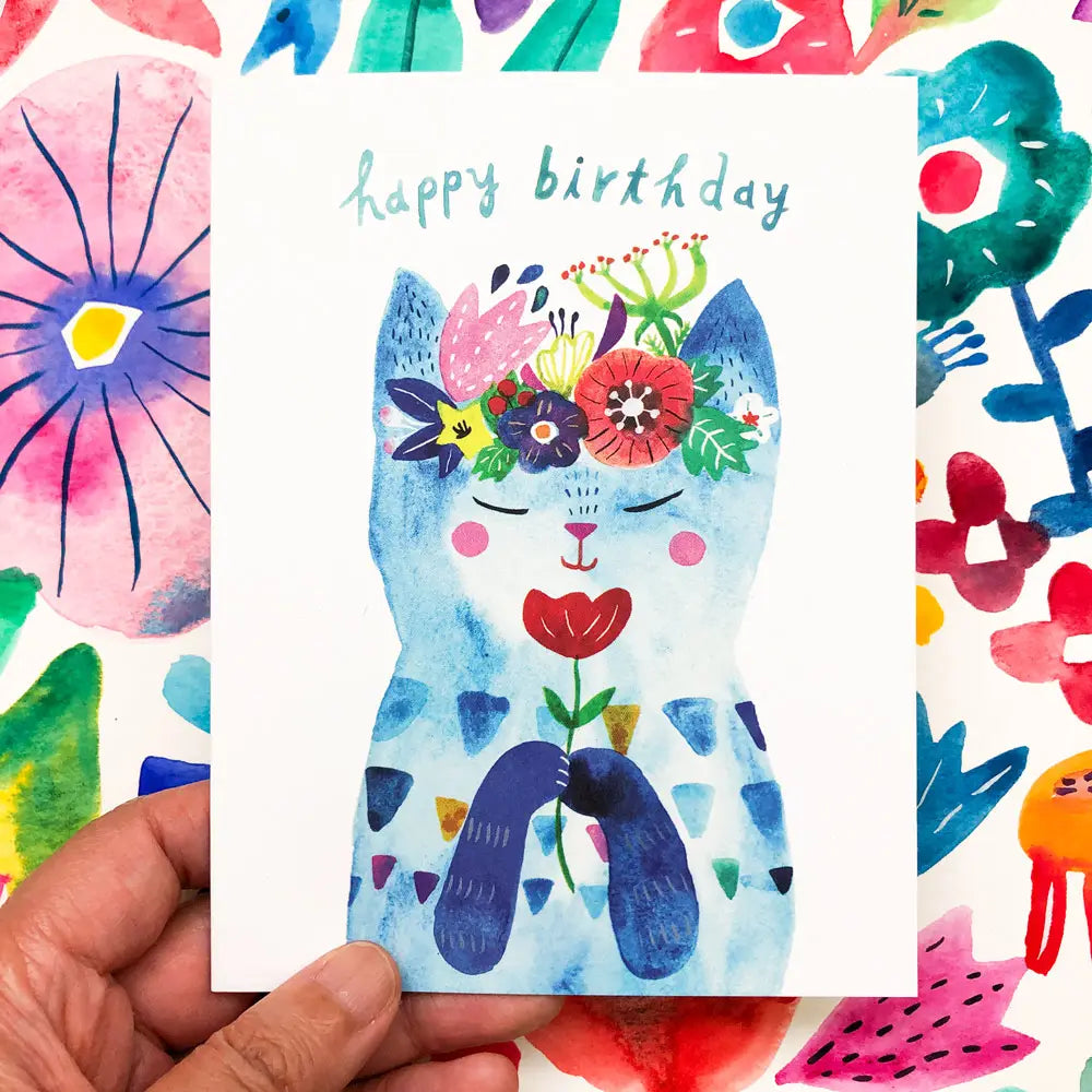 Colorful birthday card with a cat illustration and floral elements, held by a hand.