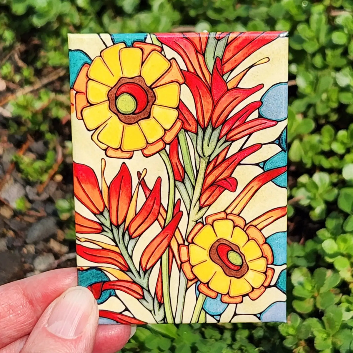 Colorful indian paintbrush design on a small rectangular magnet held in front of green foliage.
