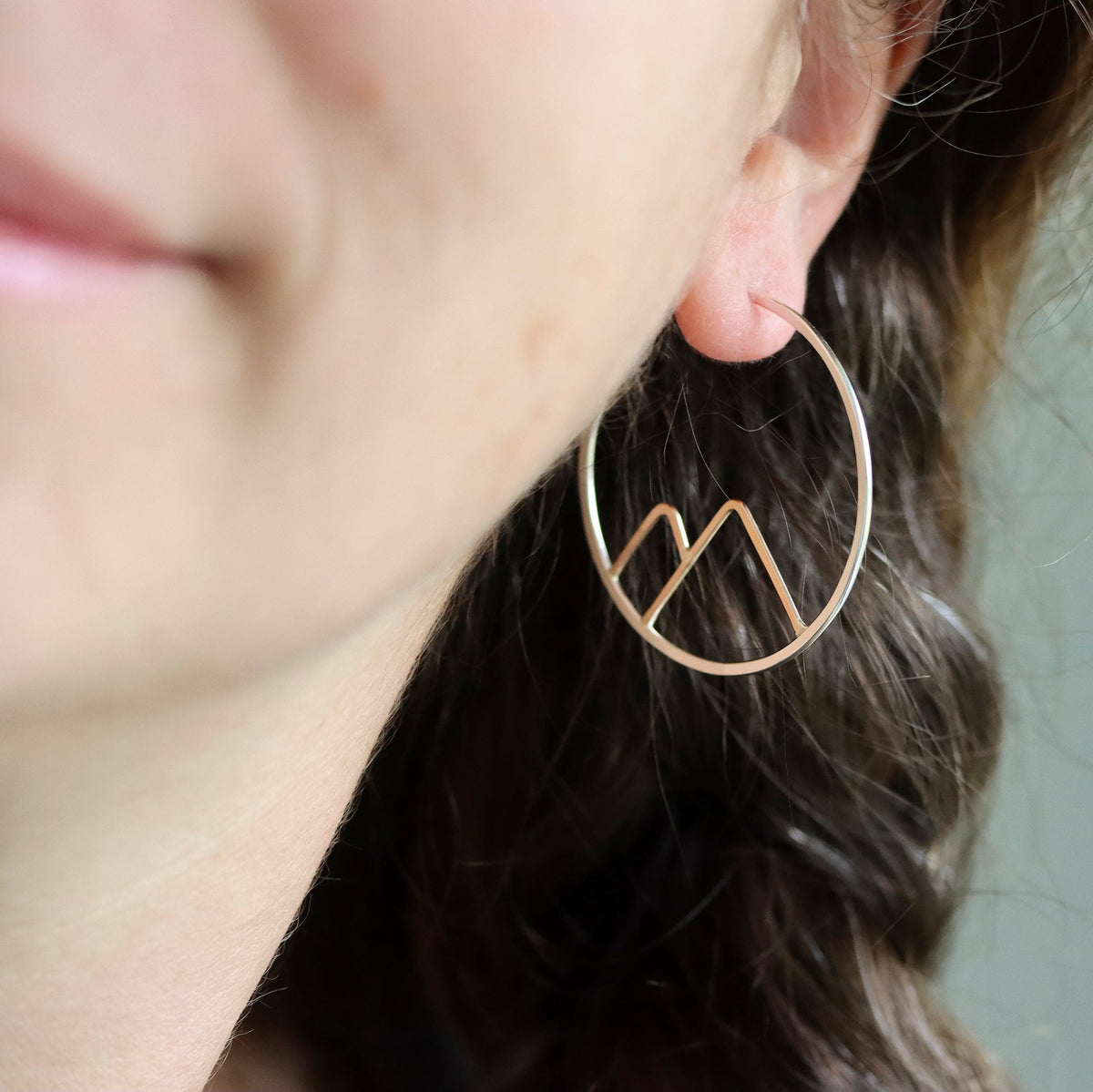 Close-up of a person wearing a gold hoop earring with a mountain design.