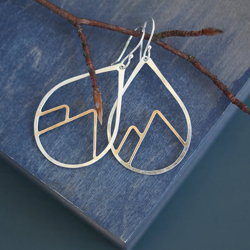 Silver mountain-shaped earrings on a blue wooden surface with a branch.