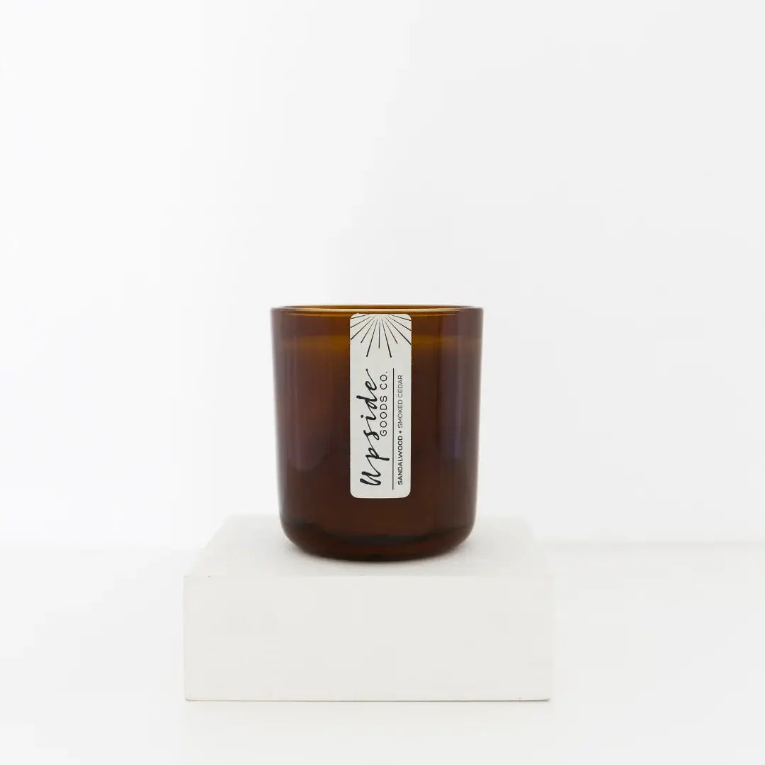 An amber glass jar candle with a white paper label reading 'upside goods' and 'sandalwood + smoked cedar'  displayed on a white pedestal against a white background.