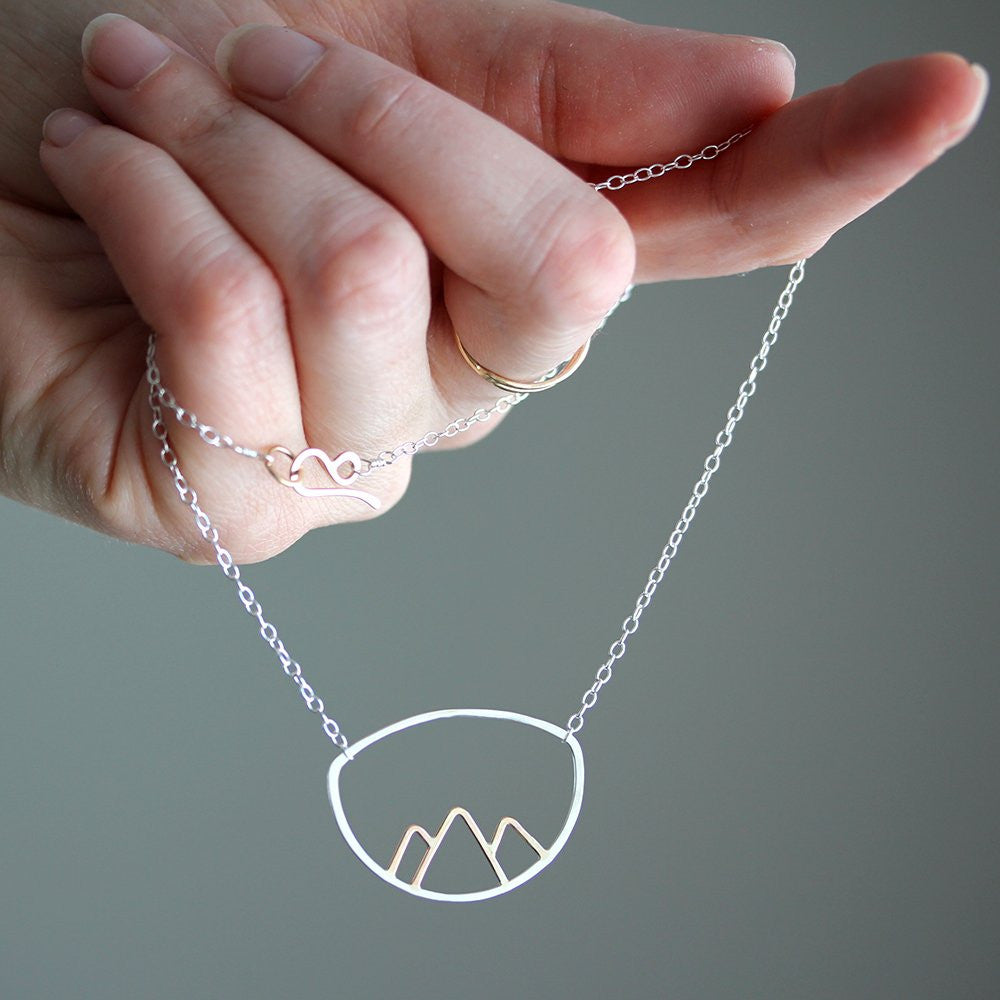 Hand holding a necklace with a mountain design against a gray background