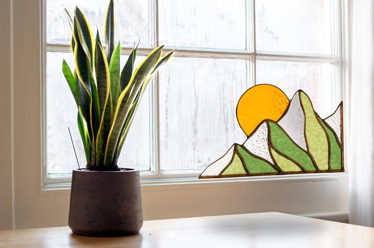 Potted plant on a windowsill with a stained glass panel of mountains and sun.