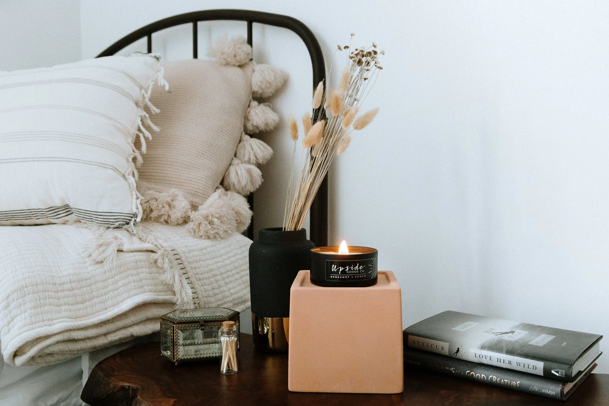 A lit candle in a black tin sits on a peach-colored block on a wooden table, styled in a cozy bedroom setting next to a bed with cream bedding and decorative pillows.