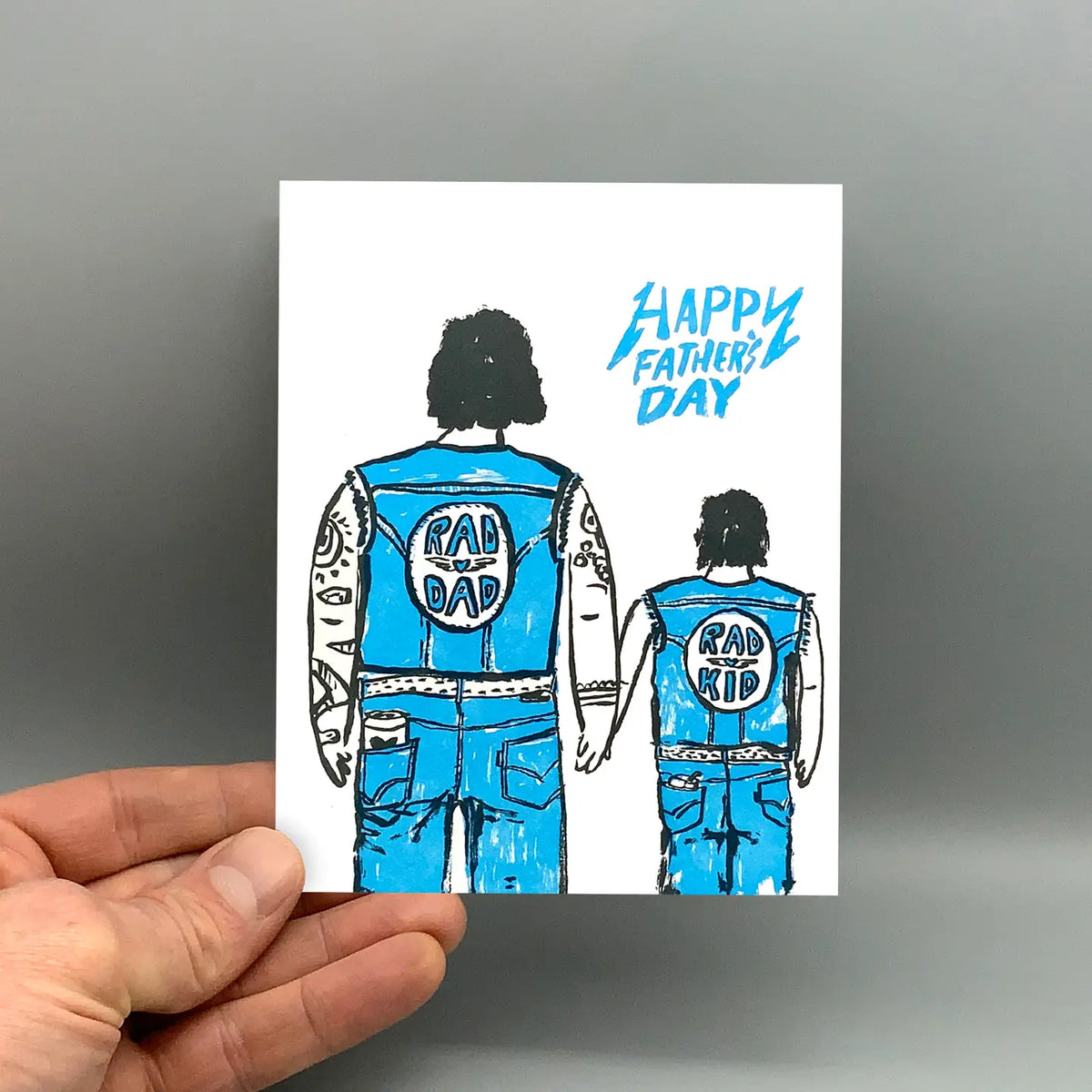 Hand holding a Father's Day card with illustrations of a father and son in blue outfits.