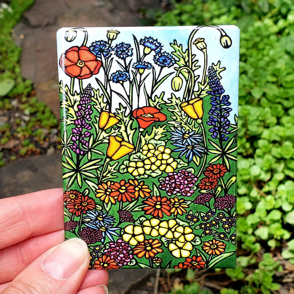 Colorful floral design on a small rectangular magnet held in front of green plants.