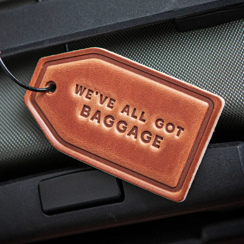 Brown leather luggage tag with 'WE'VE ALL GOT BAGGAGE' text on a car interior background