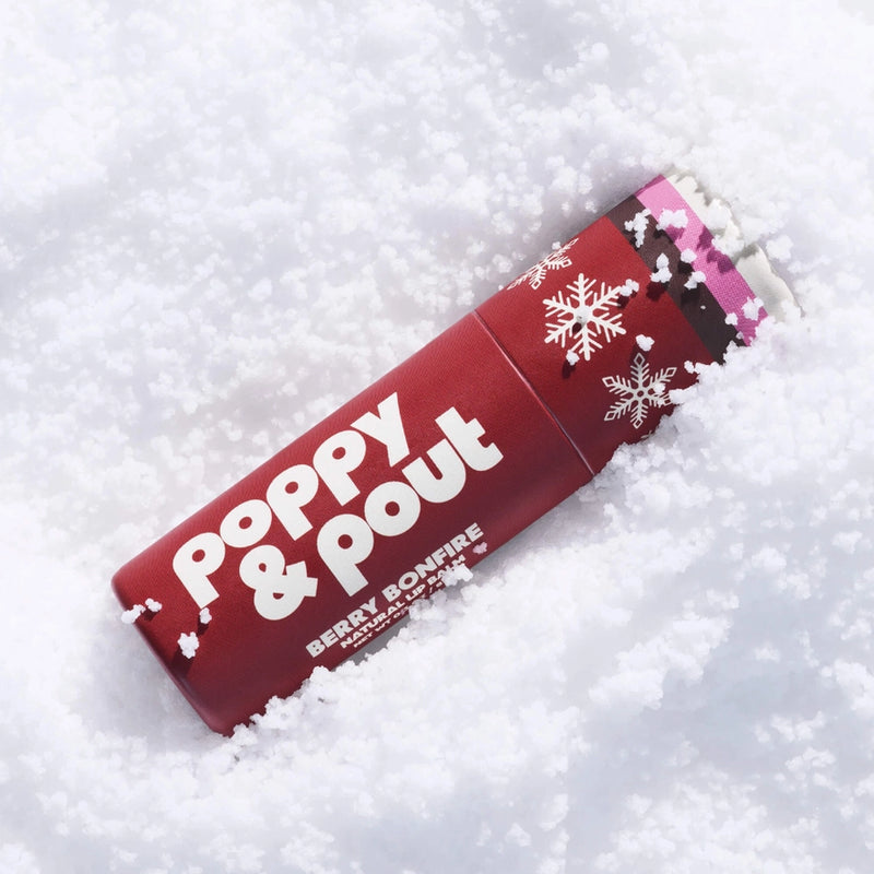 A red cylindrical container of Poppy & Pout Berry Bonfire Popcorn in the snow, with white snowflake graphics and white branding text.
