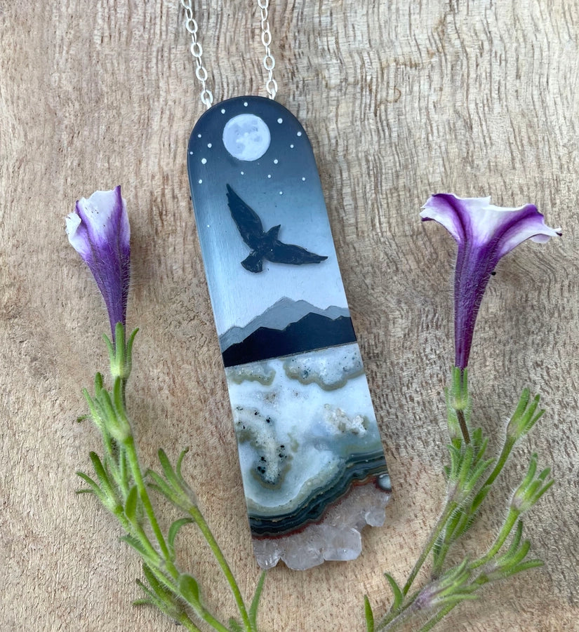 Necklace with pendant featuring a bird and mountain design, surrounded by purple flowers on a wooden surface.
