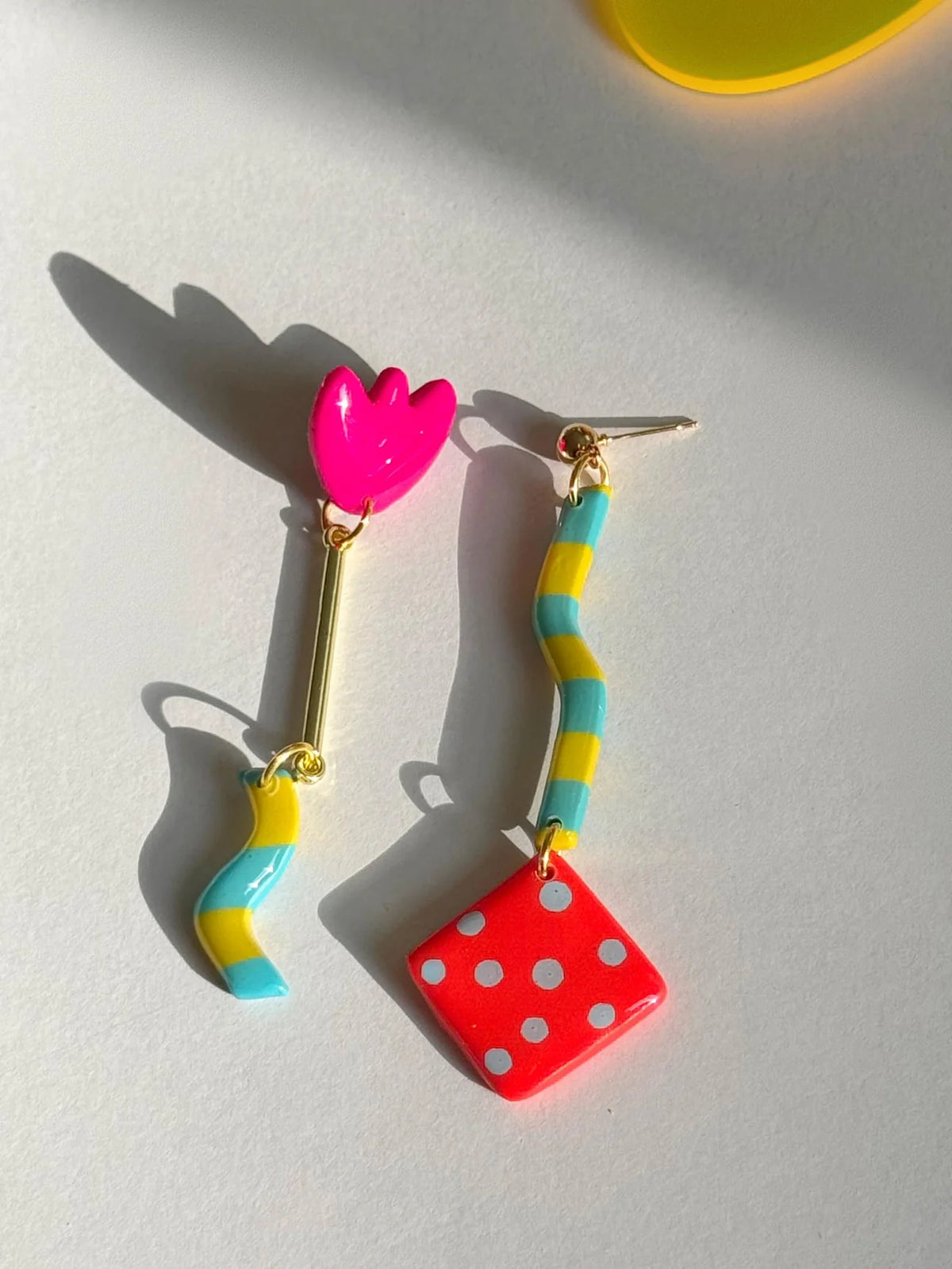 A pair of colorful dangling earrings on a light gray surface with strong sunlight and shadows. One earring has a bright pink tulip-shaped charm with a gold stem and a yellow-and-blue striped lower charm.