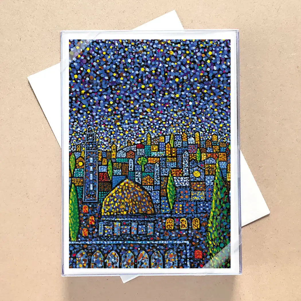 A square art print or card featuring a colorful, textured mosaic-style cityscape under a starry night sky, displayed on top of a white envelope against a beige background.