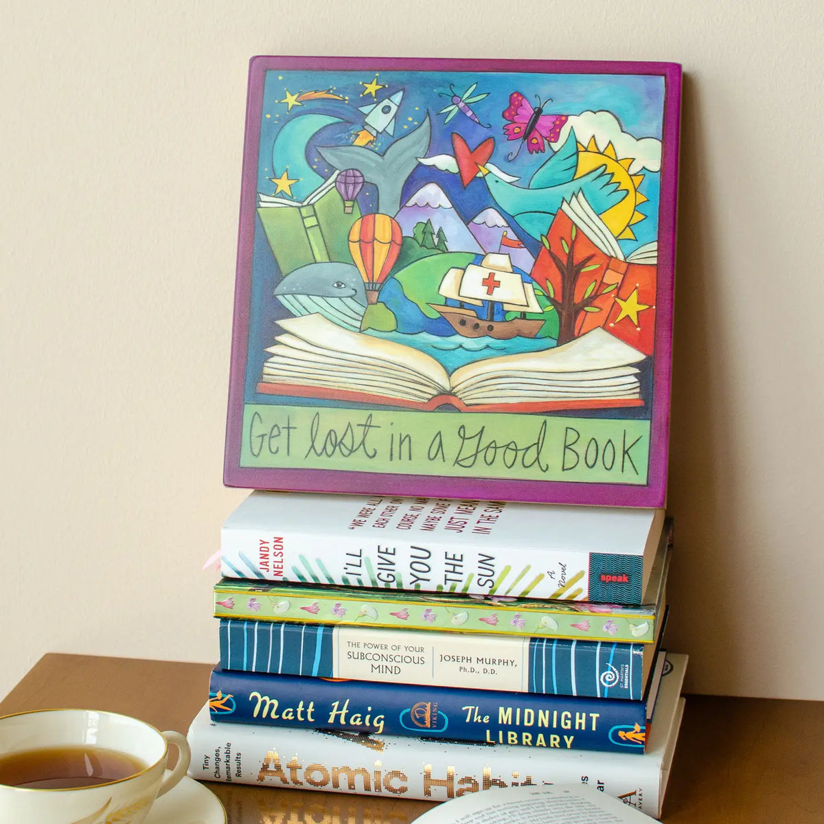 Stack of books with a colorful illustration on top and a cup of tea on a wooden surface.