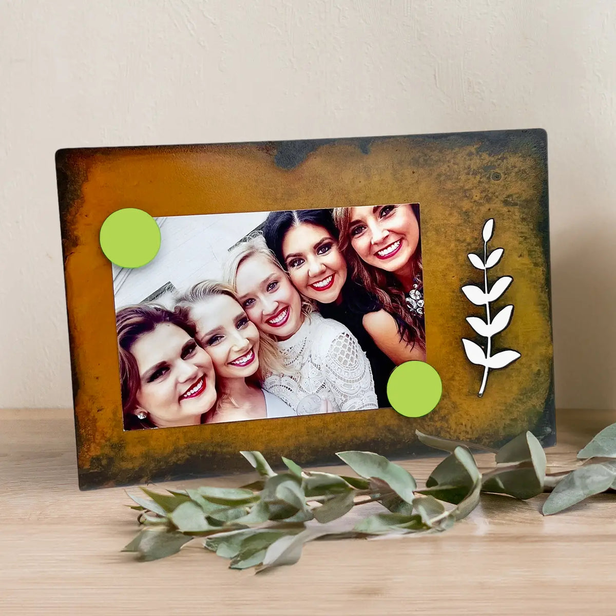 A rectangular photo frame with a distressed brown and gold finish, displayed on a light wood surface against a beige wall. The frame features two bright green circular accents, a white leaf motif on the right side, and a sample photo of four smiling women inserted on the left.