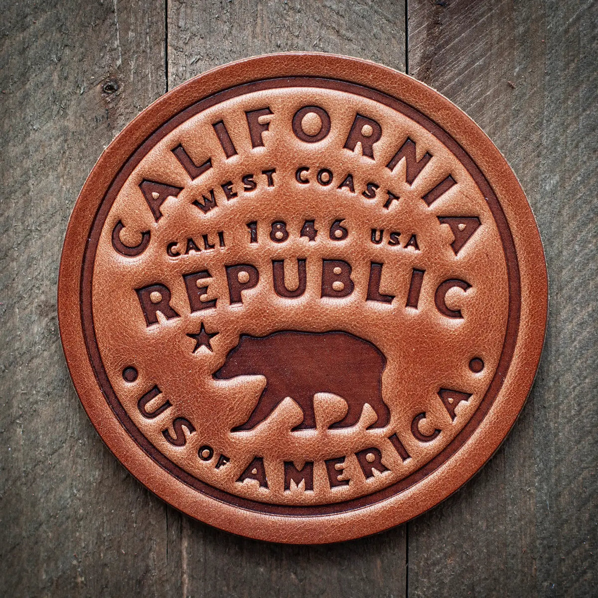 A round brown leather coaster featuring an embossed California Republic design with a bear silhouette and text, displayed on a rustic wooden surface.