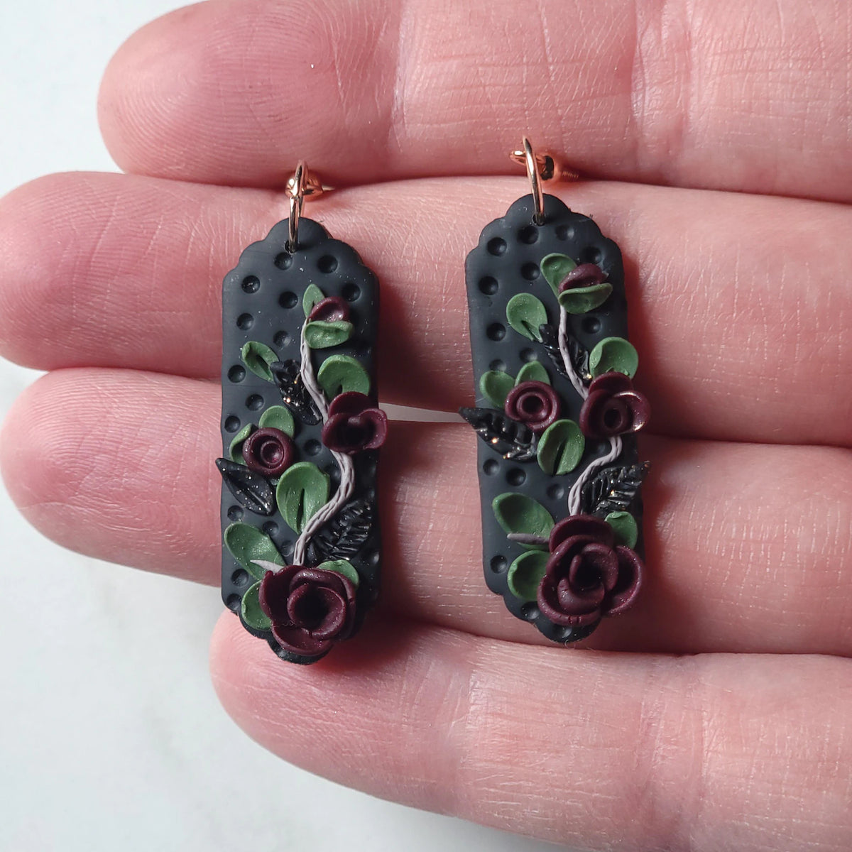 A pair of handmade polymer clay dangle earrings held in a person's hand. The earrings feature a textured black base with raised green leaves and burgundy roses, suspended from rose gold hooks.