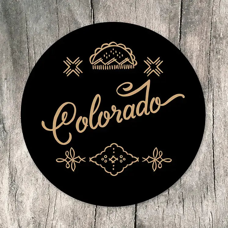 A round black sticker featuring the word 'Colorado' in elegant gold cursive script, surrounded by decorative gold line art including a mountain silhouette, stars, and ornamental flourishes, displayed on a rustic gray wood background.