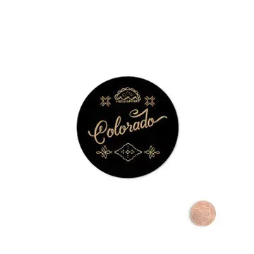 A round black sticker with gold foil-style lettering that reads 'Colorado' and decorative mountain motifs, shown on a white background with a penny for scale.
