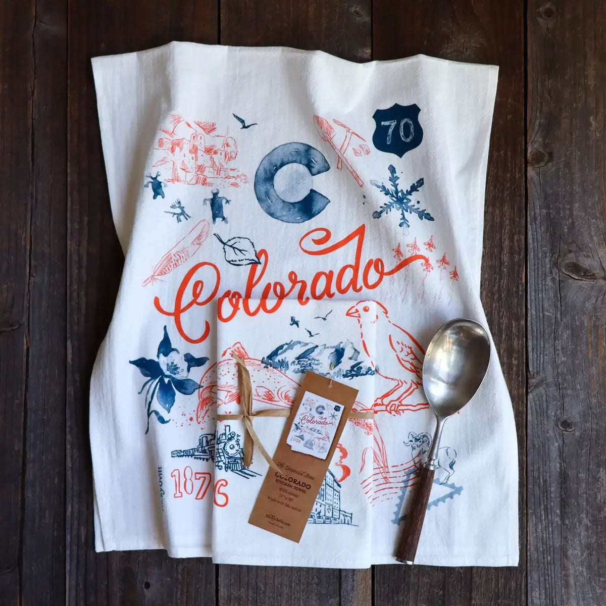 A white tea towel with a colorful Colorado-themed graphic print, laid flat on a dark wooden surface alongside a metal spoon and a product tag.