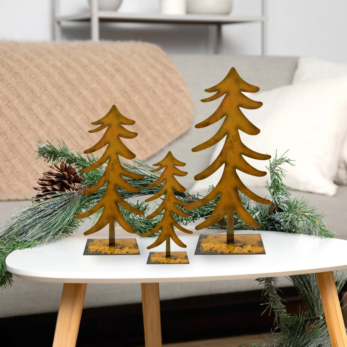 Three rusted metal Christmas tree cutouts on small square bases, displayed on a white table with evergreen garland and a pinecone in a living room setting.
