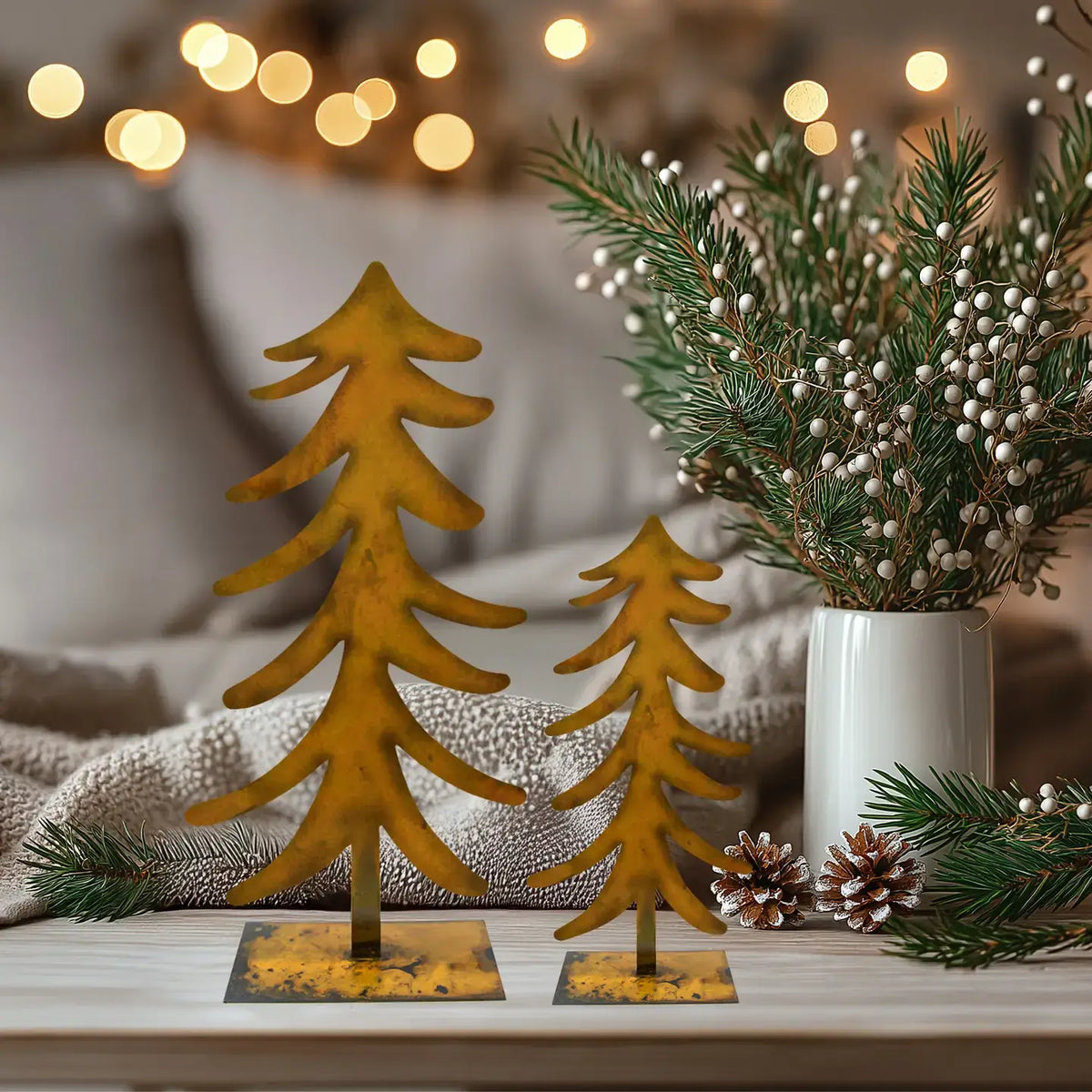 Two rustic metal Christmas tree ornaments in a warm mustard-gold finish, displayed on a light wood surface next to a white vase with green pine branches and berries, with a cozy, blurred background featuring warm bokeh lights.