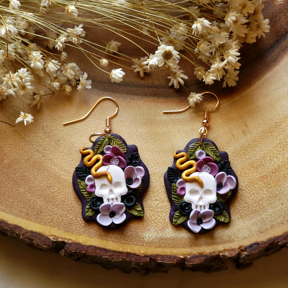 A pair of gothic-style polymer clay dangle earrings featuring white skulls, purple flowers, green leaves, and a yellow snake on a dark purple background, displayed on a rustic wood slice with dried white flowers.