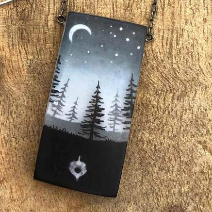 Necklace with a pendant featuring a night sky with trees and a moon on a wooden surface