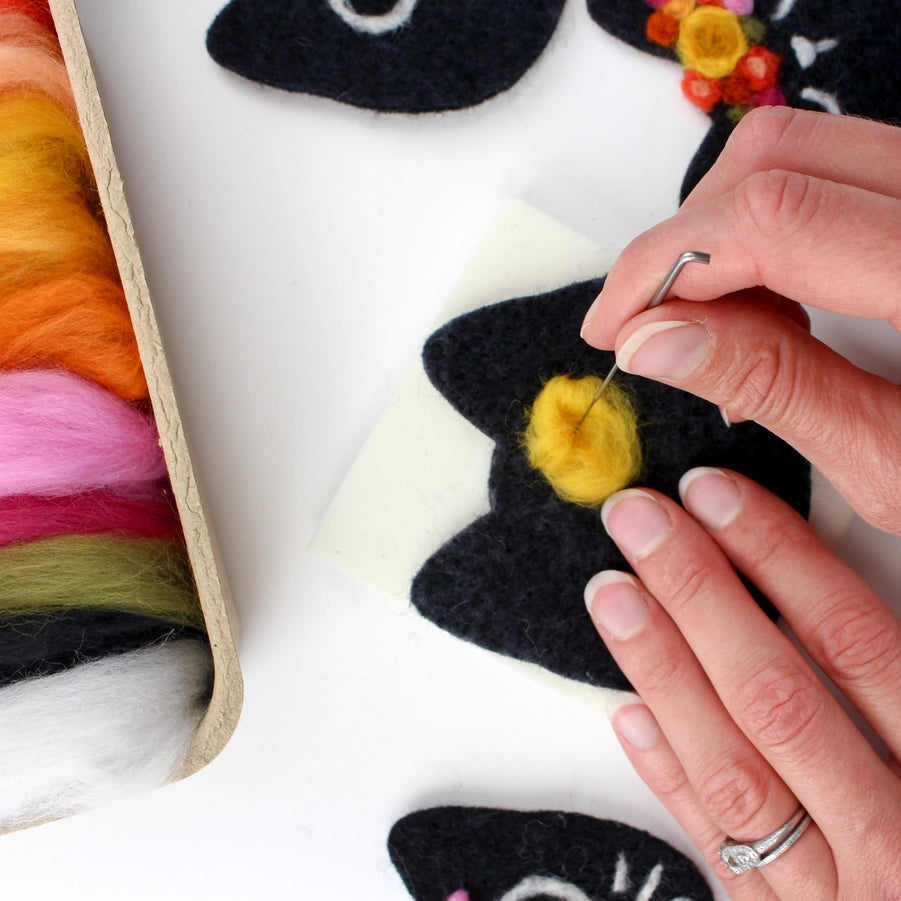 Person felting a black cat with yellow center on a white surface with colorful fibers nearby.