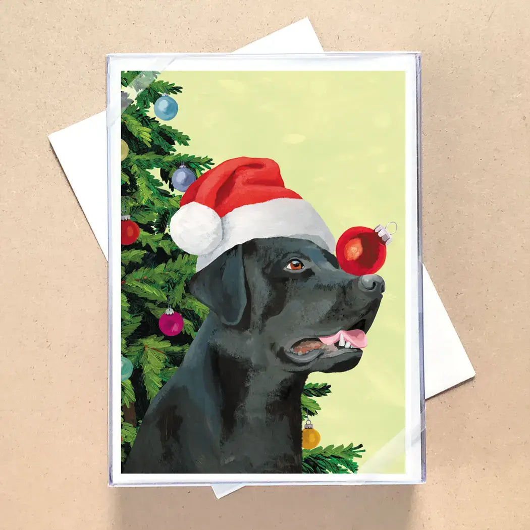 A square greeting card featuring a painted black dog wearing a red Santa hat beside a decorated Christmas tree, shown with a white envelope on a tan textured background.
