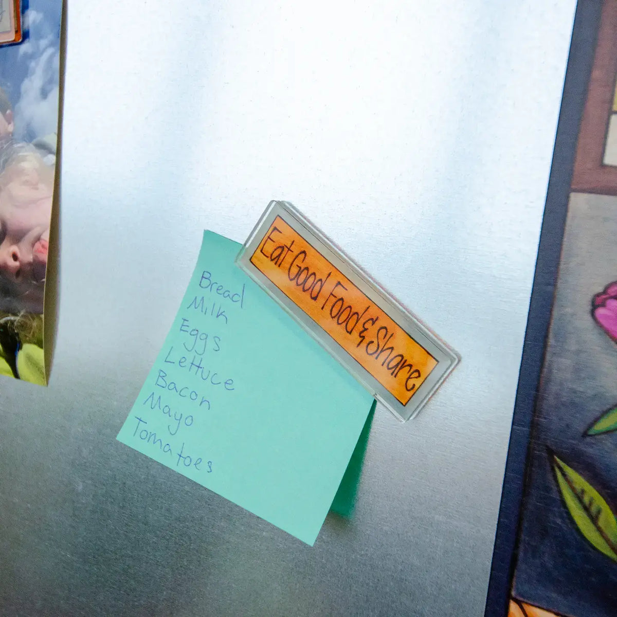 A rectangular orange magnet  with black text reading 'Eat Good Food & Share' is attached to a stainless steel refrigerator door, next to a light blue sticky note listing ingredients.