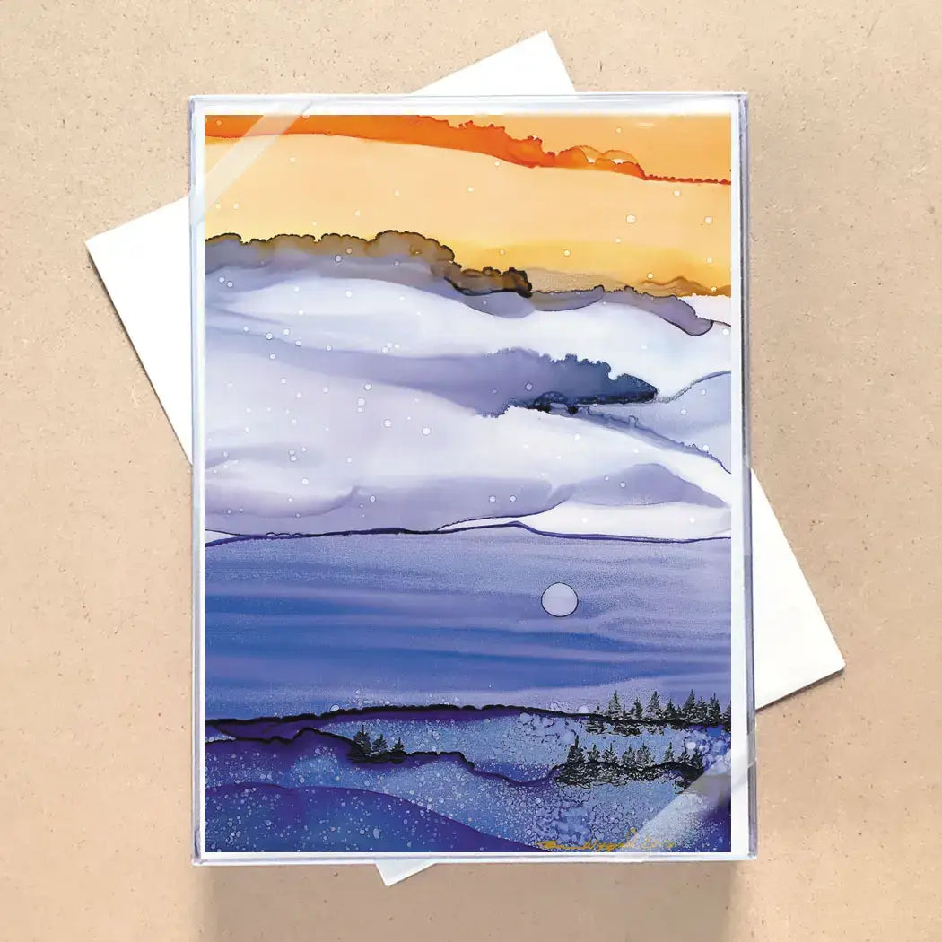 A vertical art print or greeting card featuring a watercolor landscape with snowy white and blue hills under an orange sky, shown standing on top of white envelopes against a tan background.