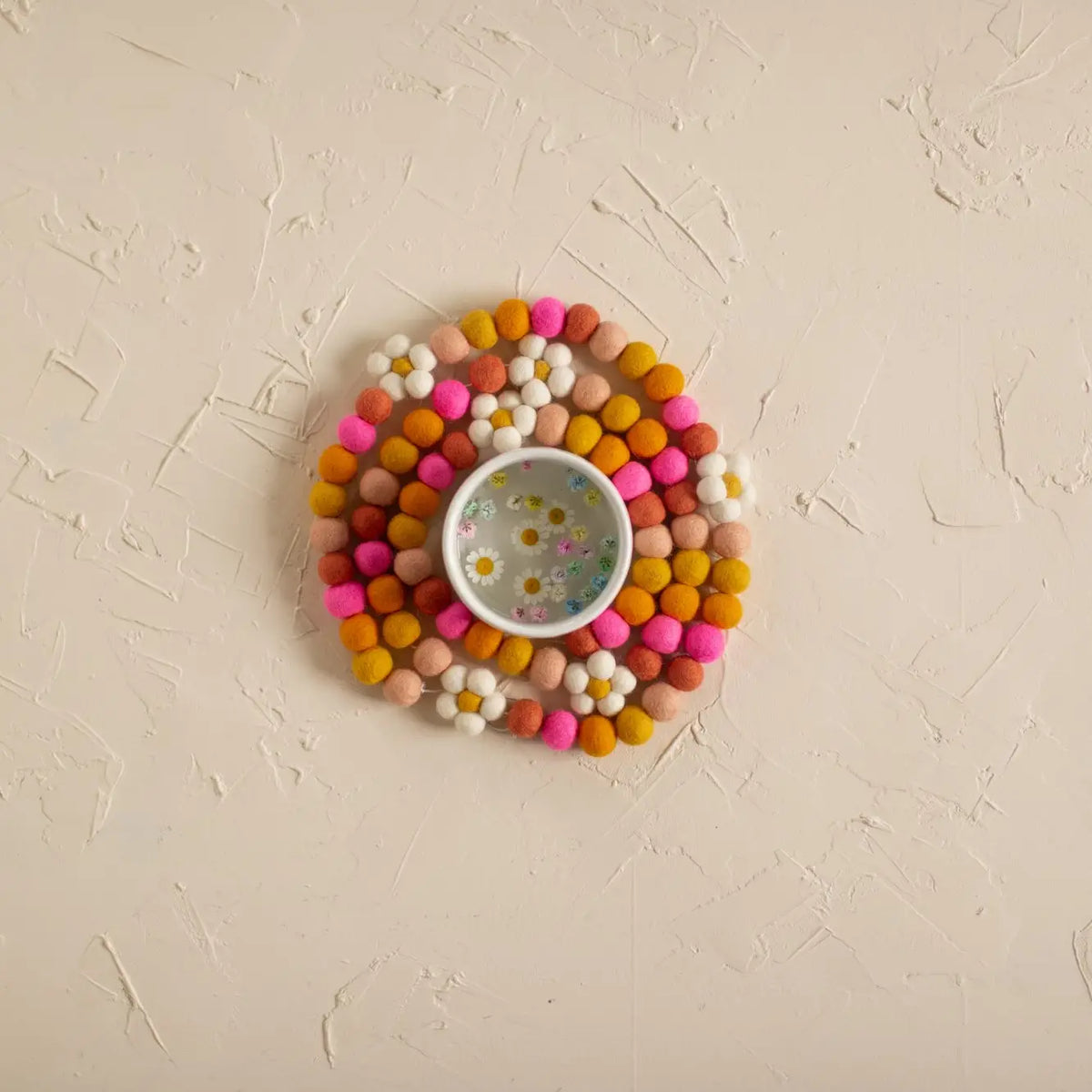 Round colorful pom-pom mat with a white bowl on a beige textured surface