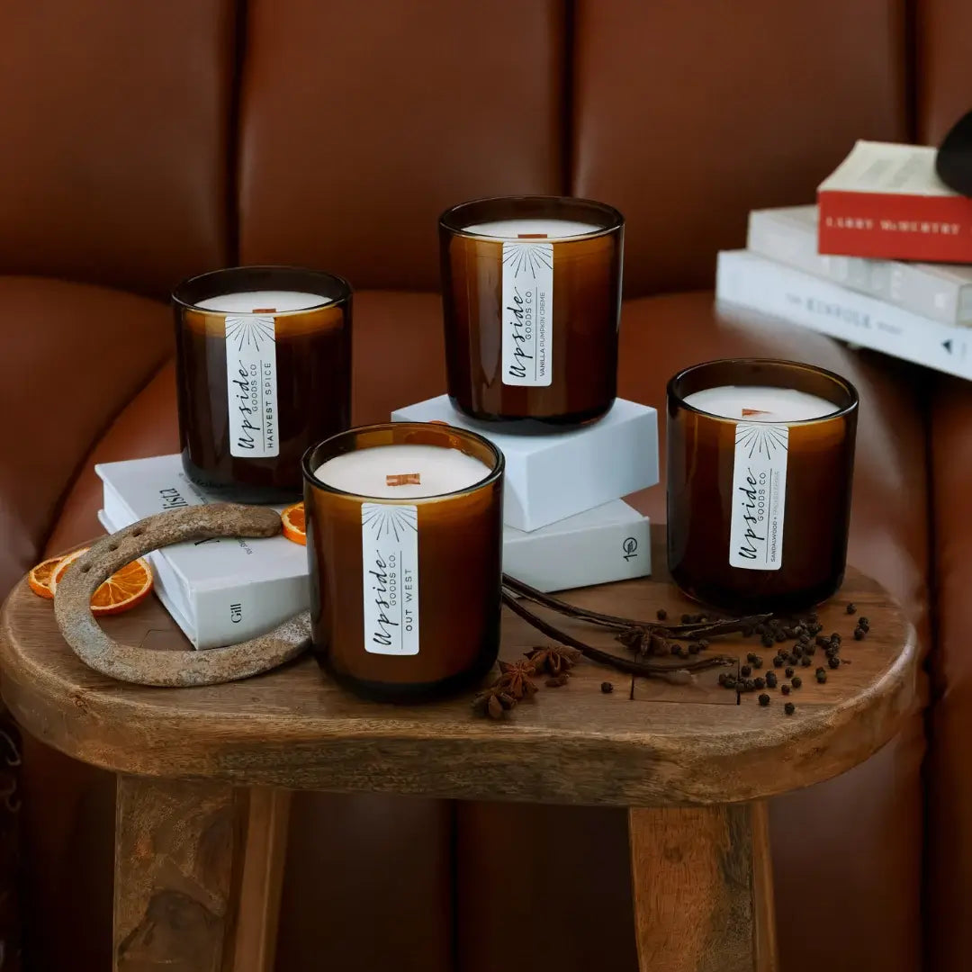 Four amber glass scented candles with white labels are arranged on a wooden stool in a warm brown setting, with books and dried orange slices as props.