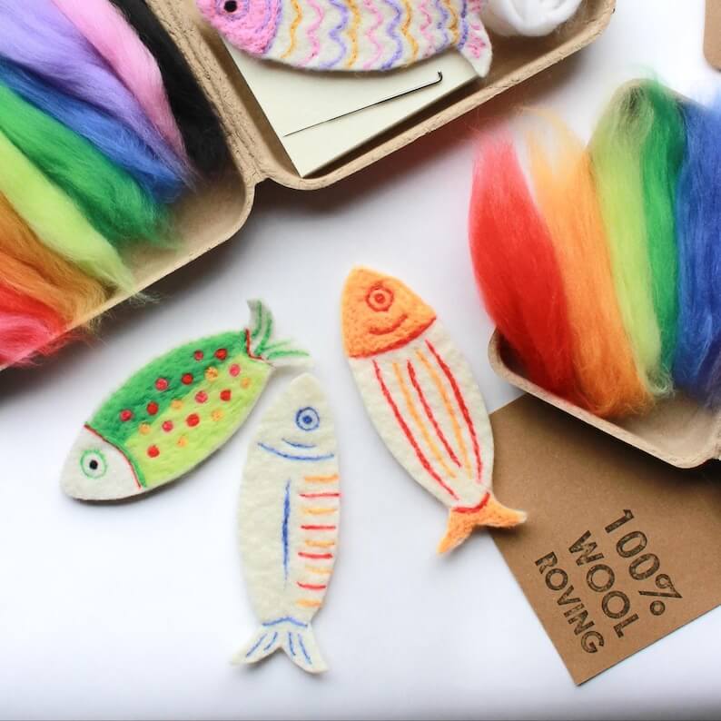 Three colorful fish-shaped felt ornaments displayed on a white surface, with open cardboard boxes of rainbow wool fibers and a tag reading '100% WOOL ROVING' in the background.