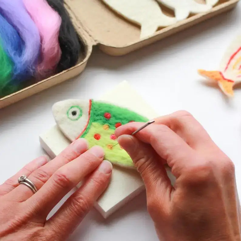 A close-up of hands needle felting a small fish-shaped piece on a white foam block, with colorful wool fibers in a box and another finished fish visible in the background.