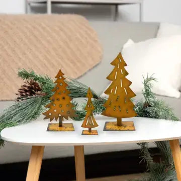 Three rust-colored metal Christmas tree cutouts on small square bases, displayed on a white table with pine branches and a blanket in the background.